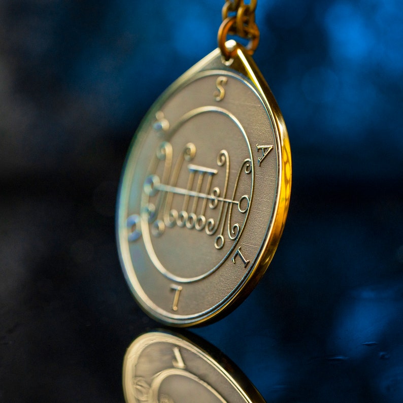 May include: A gold pendant necklace with a circular charm featuring a sigil and the letters "S", "A", "T", and "I".