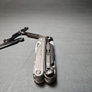 Può includere: Un multiutensile argentato con dettagli neri e la scritta "Leatherman Charge" sul lato. Lo strumento è chiuso e presenta una varietà di strumenti visibili, tra cui una lama da coltello, una pinza e un cacciavite.