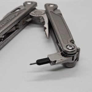 Può includere: Un multiutensile argentato con un manico nero e un piccolo accessorio per cacciavite. Lo strumento è parzialmente aperto, rivelando le pinze e altri strumenti.