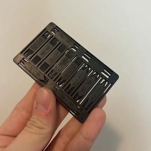 Puede incluir: Un juego de puntas de destornillador negras en un soporte rectangular. Las puntas están etiquetadas con información de tamaño. El soporte se sostiene en una mano sobre un fondo blanco. Las puntas son para varios tipos de tornillos.