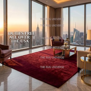 Deep wine color solid area rug displayed in a luxury modern living room with large windows and city views. Plush handcrafted rug with rich burgundy tone adds warmth and elegance to contemporary interior decor. Perfect for living room, bedroom, or apartment styling, this handmade rug features a smooth, soft texture and premium finish, ideal for modern, minimalist, and luxury home interiors.