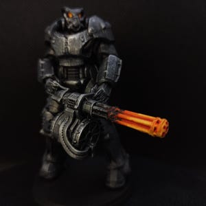 May include: A miniature figurine of a soldier in dark gray armor, holding a large machine gun with orange and yellow glowing barrels. The figure has a helmet with glowing orange eyes. The background is a solid black.
