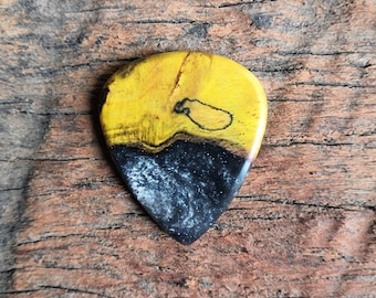May include: A guitar pick with a yellow and black design. The top half is yellow with dark wood grain and a black marking. The bottom half is black with a speckled, glittery appearance. The pick is on a wooden surface.