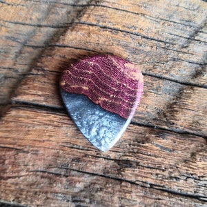 May include: A wooden guitar pick with a purple and gray wood grain pattern. The pick is shaped like a traditional guitar pick and has a smooth, polished finish.