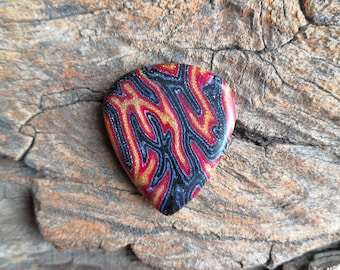 May include: A guitar pick with a black, red, and gold abstract pattern. The pick is made of a shiny material and has a pointed tip.