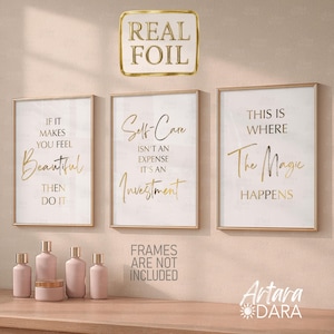 May include: Three framed wall art prints with gold foil lettering on a white background. The prints read: "If it makes you feel beautiful, then do it," "Self-Care isn't an expense, it's an investment," and "This is where the magic happens." Pink cosmetic bottles are on a wooden shelf.