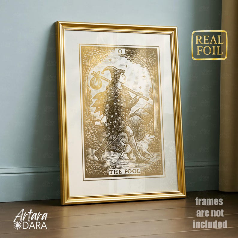 May include: A framed gold foil print of "The Fool" tarot card. The artwork features a figure with a staff and a dog, set in a forest scene. The print is in a gold frame and has the text "REAL FOIL". The artist's logo is in the bottom left corner.