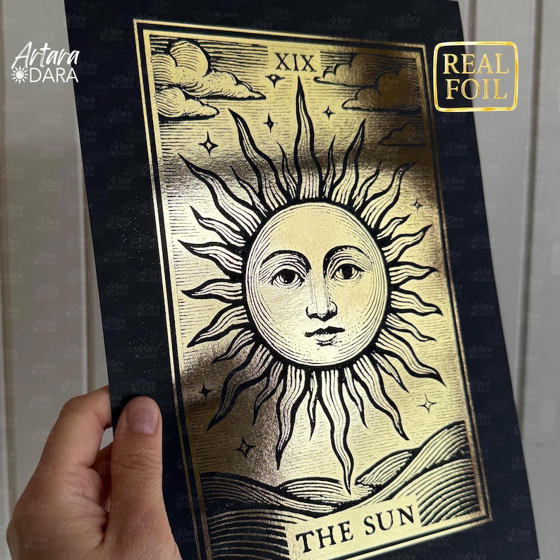May include: A gold foil tarot card print featuring the sun with a face, rays, and the text "THE SUN". The card has a black border and the number XIX at the top. The words "REAL FOIL" are in a gold square.