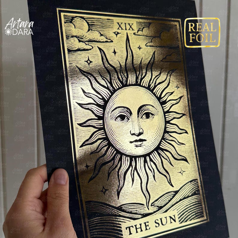 May include: A gold foil tarot card print featuring the sun with a face, rays, and the text "THE SUN". The card has a black border and the number XIX at the top. The words "REAL FOIL" are in a gold box.