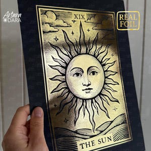 May include: A gold foil tarot card print featuring the sun with a face, rays, and the text "THE SUN". The card has a black border and the number XIX at the top. The words "REAL FOIL" are in a gold square.