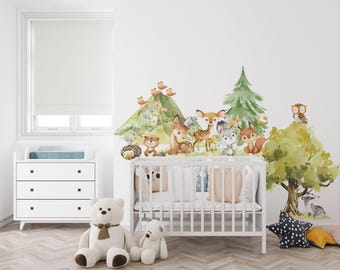 May include: A set of watercolor wall decals featuring a green mountain, a pine tree, a deciduous tree, and various forest animals including a deer, squirrel, owl, hedgehog, and rabbit, arranged on a white wall above a white wooden baby crib.
