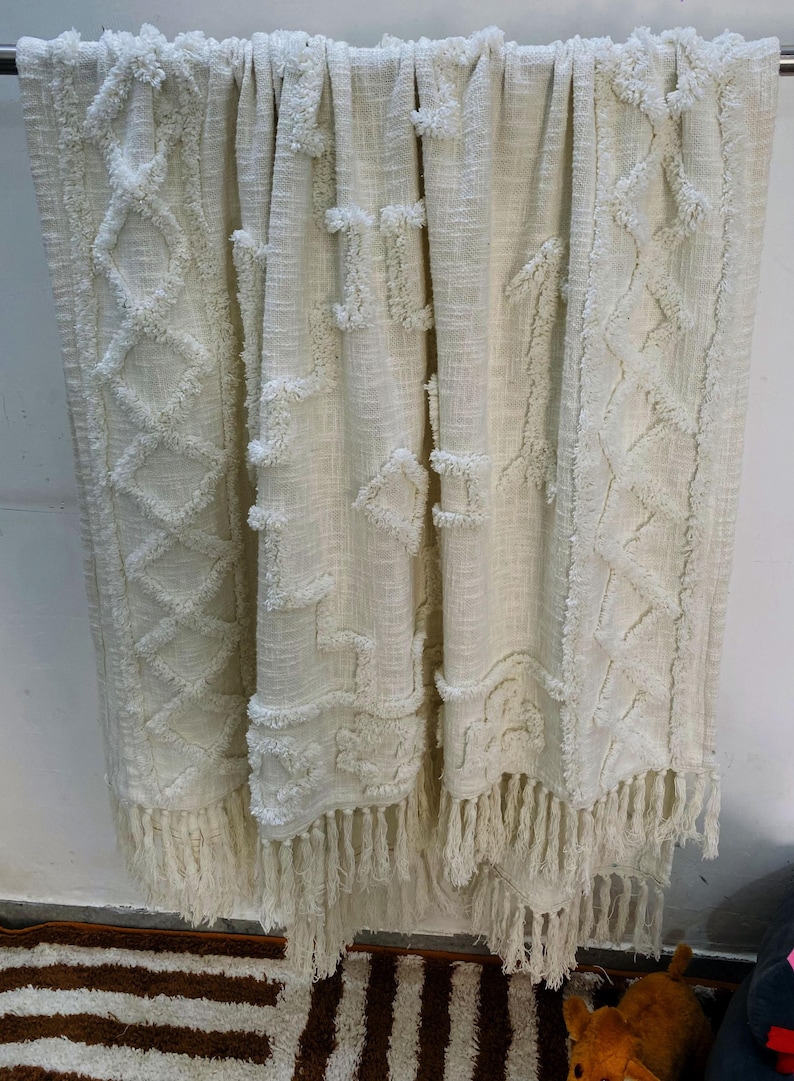 May include: Cream-coloured woven throw blanket with textured geometric patterns and fringed trim. The blanket features diamond and square designs, creating a cosy and decorative accent for any living space.