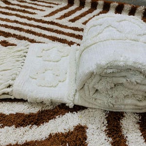 May include: A rolled-up, cream-coloured textured blanket with a fringed edge and a floral pattern. The blanket rests on a brown and white striped rug, creating a cosy scene. The rug's pattern is reminiscent of zebra stripes.