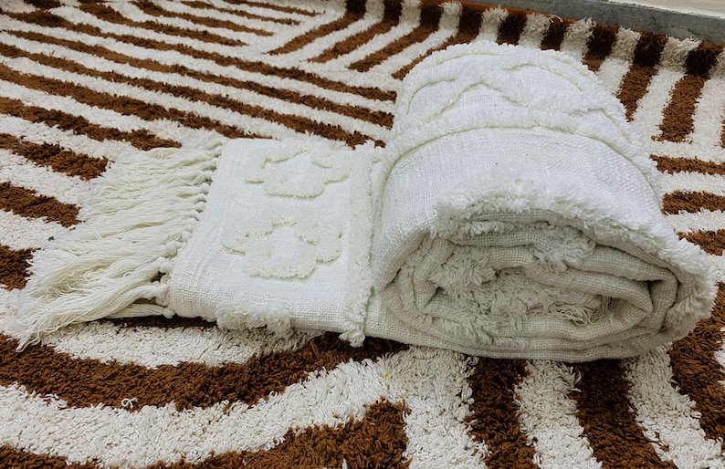 May include: A rolled-up, cream-coloured textured blanket with a fringed edge and a floral pattern. The blanket rests on a brown and white striped rug, creating a cosy scene. The rug's pattern is reminiscent of zebra stripes.