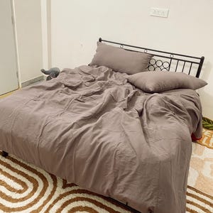 Può includere: Un letto con struttura in metallo nero e piumino e cuscini marroni. Il letto è fatto con un piumino stropicciato in tinta unita e due cuscini abbinati. Il letto poggia su un tappeto fantasia con vortici marroni e bianchi.