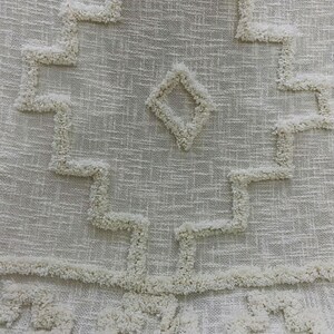 May include: Close-up of a cream-coloured textile with a geometric pattern. The design features a central diamond shape and zig-zag lines, all in a raised, textured material. The fabric has a woven appearance.