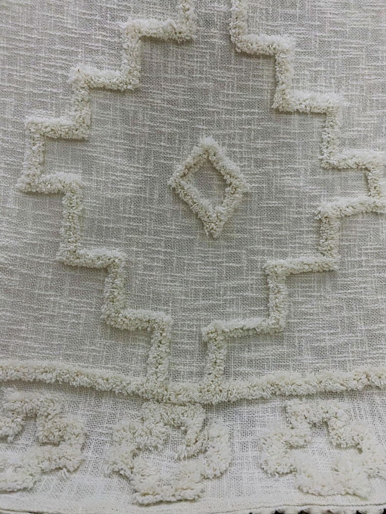 May include: Close-up of a cream-coloured textile with a geometric pattern. The design features a central diamond shape and zig-zag lines, all in a raised, textured material. The fabric has a woven appearance.