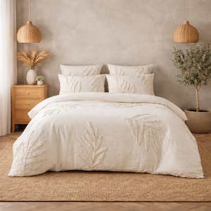 Puede incluir: Juego de cama color crema con diseños de hojas en relieve. El juego incluye una funda nórdica y fundas de almohada a juego. La decoración del dormitorio también incluye una mesita de noche de madera, lámparas colgantes tejidas y un olivo en maceta.