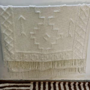 May include: Off-white woven throw blanket with geometric patterns and fringe. The blanket features diamond, arrow, and stepped designs. The bottom edge has a long fringe. The blanket is hanging against a white wall.