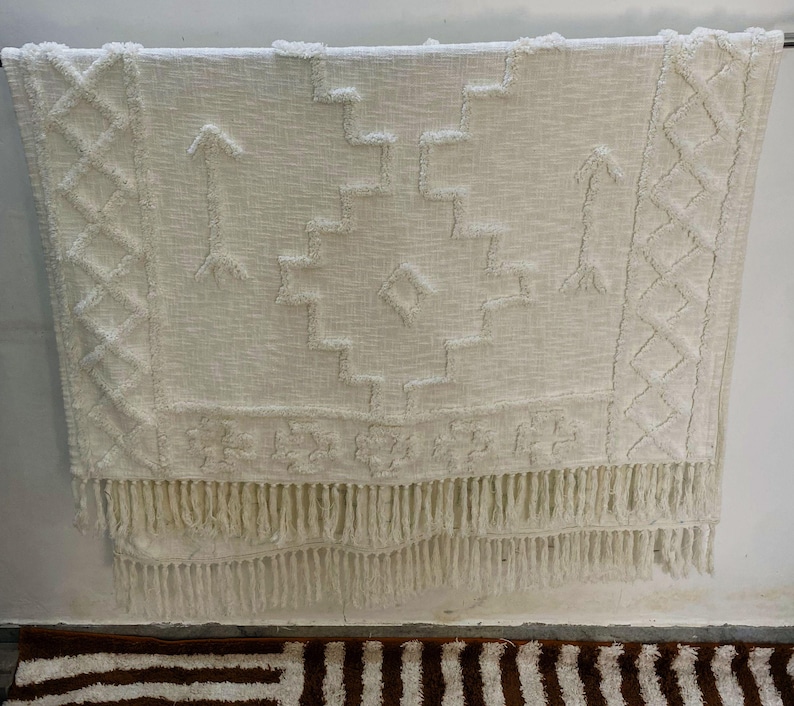 May include: Off-white woven throw blanket with geometric patterns and fringe. The blanket features diamond, arrow, and stepped designs. The bottom edge has a long fringe. The blanket is hanging against a white wall.