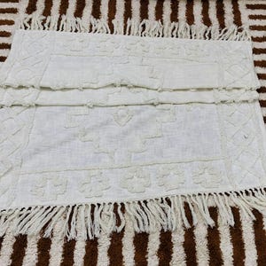 May include: Off-white rectangular rug with a textured pattern and fringe trim. The rug features a repeating floral and geometric design. The rug is placed on a brown and white striped surface.