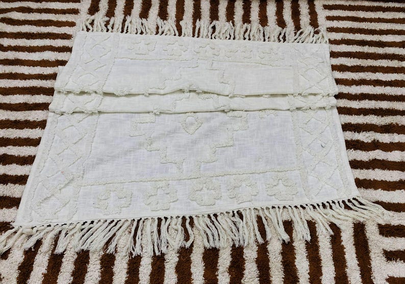 May include: Off-white rectangular rug with a textured pattern and fringe trim. The rug features a repeating floral and geometric design. The rug is placed on a brown and white striped surface.