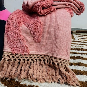 May include: A pink throw blanket with a textured design and fringe. The blanket has a woven texture with a raised, shaggy pattern in a darker shade of pink. The bottom edge features a long fringe.