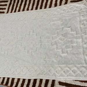 May include: Off-white rectangular rug with a geometric pattern and fringe trim. The rug features a textured design with diamond and step-like shapes. The rug is placed on a brown and white patterned rug.
