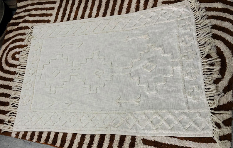 May include: Off-white rectangular rug with a geometric pattern and fringe trim. The rug features a textured design with diamond and step-like shapes. The rug is placed on a brown and white patterned rug.