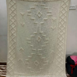 May include: Cream-coloured woven throw blanket with a geometric pattern and fringe trim. The blanket features diamond, arrow, and rectangular shapes in a textured design. The edges are finished with a fringe.