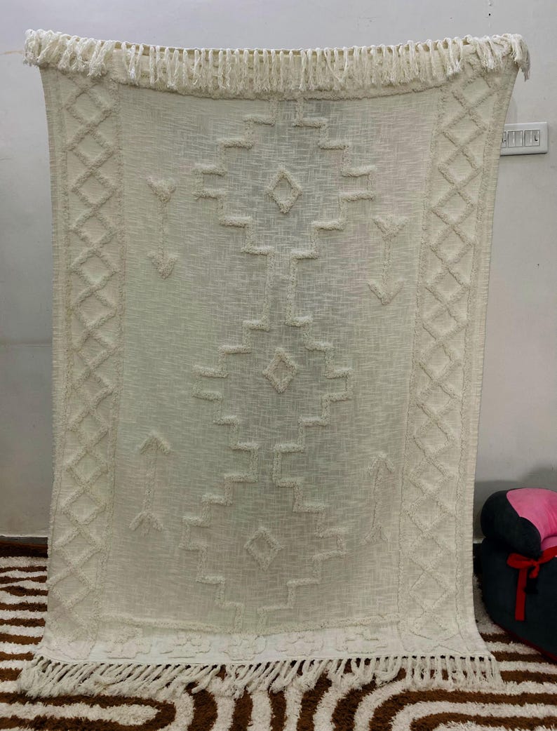 May include: Cream-coloured woven throw blanket with a geometric pattern and fringe trim. The blanket features diamond, arrow, and rectangular shapes in a textured design. The edges are finished with a fringe.