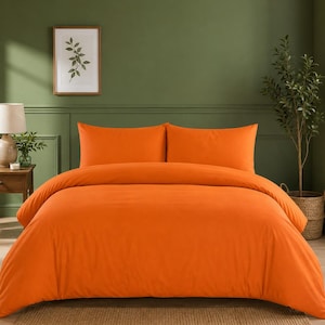 May include: An orange bed set, including a duvet cover and two pillowcases, is displayed on a bed. The bedding is the focal point, with a solid, vibrant color. The background features a green wall, a framed botanical print, and a small potted plant.