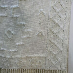May include: Off-white woven rug with geometric patterns and fringe. The rug features raised, textured designs including diamonds, arrows, and stepped shapes. The bottom edge has a long fringe.