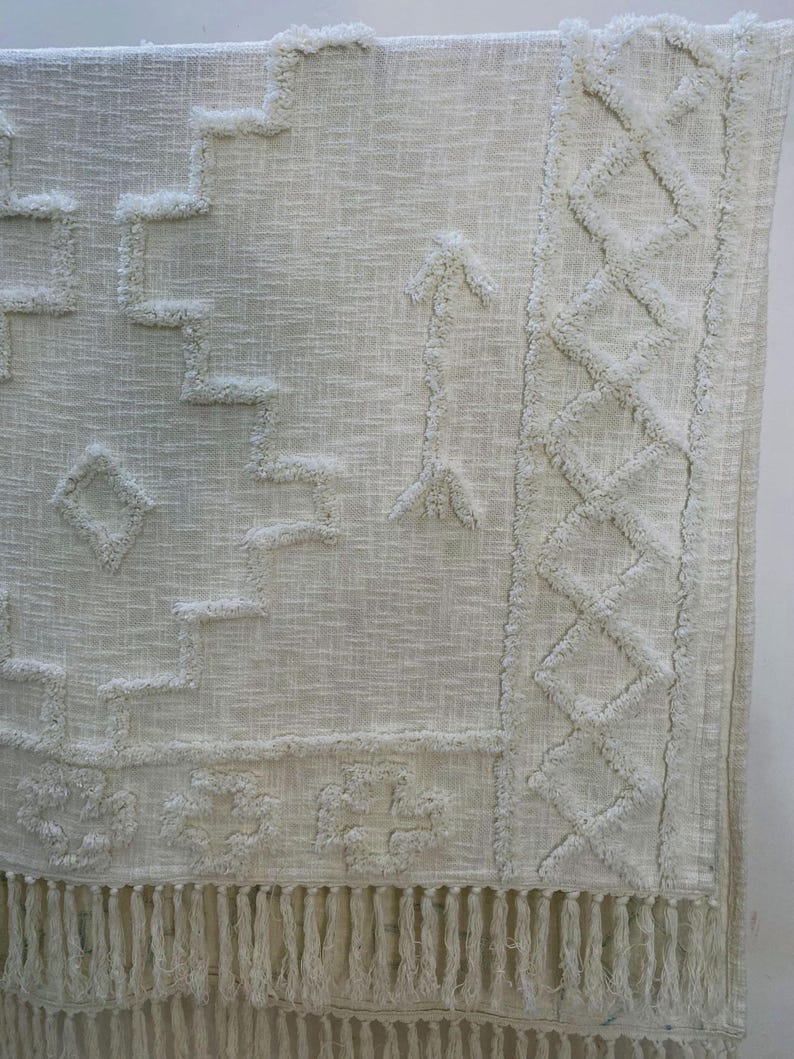 May include: Off-white woven rug with geometric patterns and fringe. The rug features raised, textured designs including diamonds, arrows, and stepped shapes. The bottom edge has a long fringe.