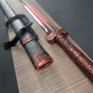 May include: A decorative sword with a red and black scabbard. The scabbard has intricate carvings and a tassel. The sword has a red and gold handle with intricate carvings.