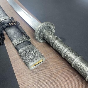 May include: A silver-coloured sword with a black scabbard. The scabbard is decorated with intricate silver designs. The sword has a silver handle with a dragon design.