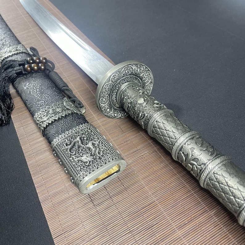 May include: A silver-coloured sword with a black scabbard. The scabbard is decorated with intricate silver designs. The sword has a silver handle with a dragon design.