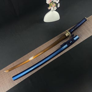 Puede incluir: Una katana, espada japonesa tradicional, con una vaina azul y dorada y una hoja de color cobre. La espada se muestra en una estera negra con un patrón de rejilla de madera.