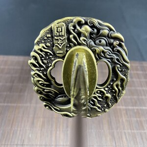 May include: A gold-coloured metal sword tsuba, or handguard, with a detailed design of a dragon's head and swirling clouds. The tsuba has two circular openings and a raised, textured centre.