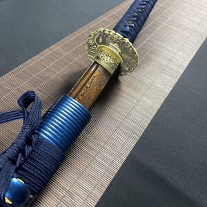 May include: A close-up of a katana sword with a blue and black handle and a gold-coloured tsuba, or hand guard, with intricate designs. The sword is resting on a brown woven mat.