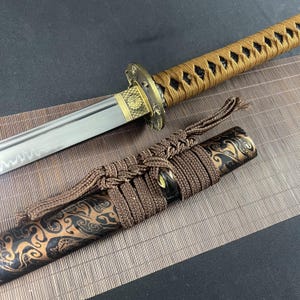 Puede incluir: Una katana japonesa tradicional con una vaina de patrón marrón y negro y un cordón trenzado marrón y negro. La espada tiene una empuñadura dorada con un diseño decorativo.