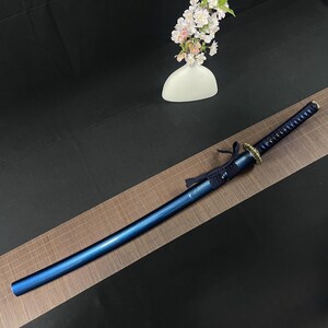 May include: A blue katana sword with a black and gold handle. The sword is lying on a brown mat with a grid pattern.