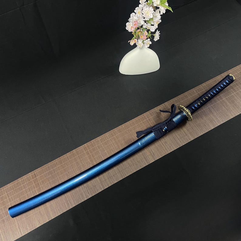 May include: A blue katana sword with a black and gold handle. The sword is lying on a brown mat with a grid pattern.