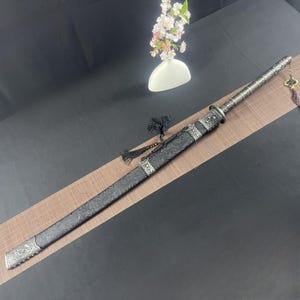 May include: A black sword with a silver-coloured hilt and scabbard. The scabbard has intricate designs and is decorated with a tassel.