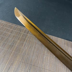May include: A close-up of the blade of a katana sword. The blade is a golden colour and has a textured surface.