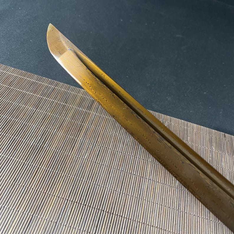 May include: A close-up of the blade of a katana sword. The blade is a golden colour and has a textured surface.