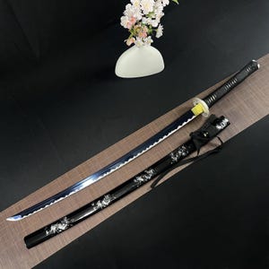 Puede incluir: Una katana negra y azul con una vaina de patrón blanco y negro. La espada tiene un mango negro con un acento amarillo.