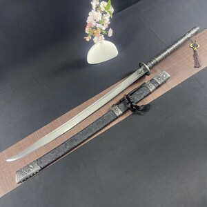 May include: A silver and black samurai sword with a decorative handle and a black scabbard with intricate designs. The sword is on a black surface with a white vase of flowers in the background.