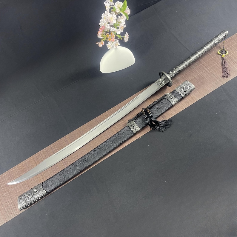 May include: A silver and black samurai sword with a decorative handle and a black scabbard with intricate designs. The sword is on a black surface with a white vase of flowers in the background.