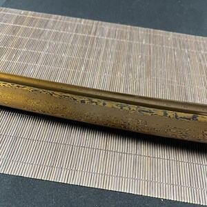 May include: A close-up of a gold and black sword blade with a wavy pattern. The blade is resting on a woven bamboo mat.